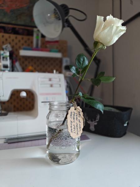 A white rose sits in a mason jar. The stem has a label affixed to it with twine. The label says "Did you know... 88% of men never receive flowers until their funeral"