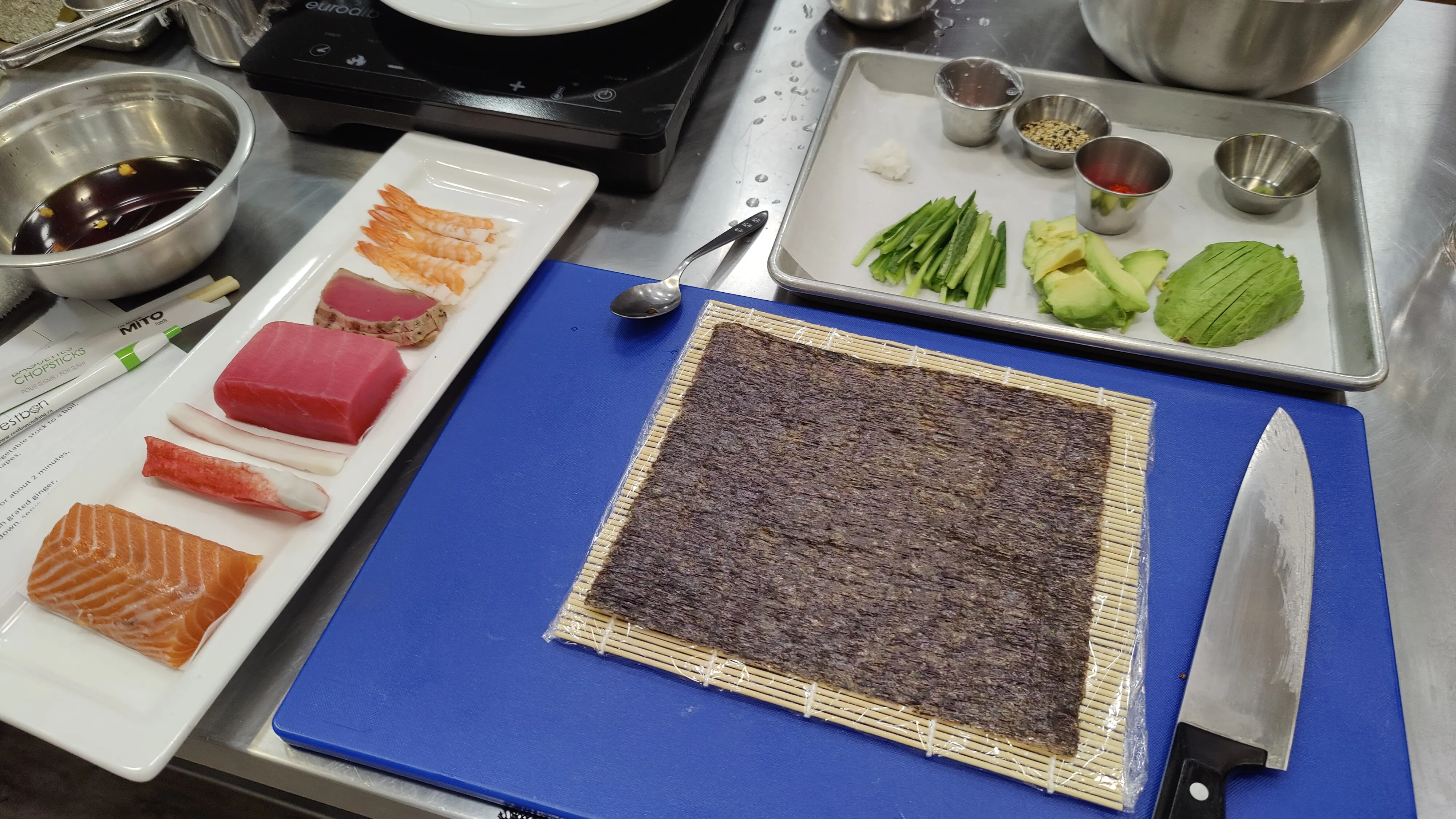 Sliced cucumber and avocado, raw salmon, tuna, crab, grilled tuna, and shrimp sit on a tray next to a cutting board, which holds a bamboo rolling mat and a sheet of nori.